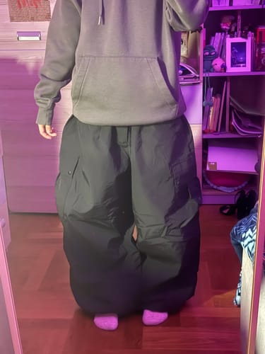 Reviewer tries on the black baggy snow pants, showing their oversized fit in a room.