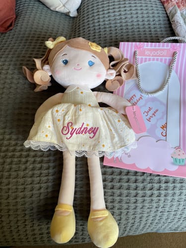 Customer's Personalized Daisy Girl doll in a yellow dress embroidered with the name 'Sydney', shown with its gift bag.