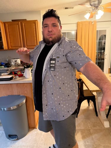 Reviewer trying on the gray Star Wars™ “Lil Troopers” shirt, worn open over a black t-shirt in a kitchen.