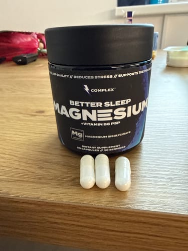 Customer's black bottle of Complex Better Sleep Magnesium with three white capsules on a wooden table.
