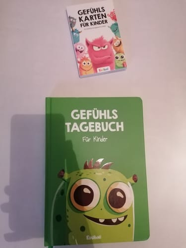 Ein Kunde zeigt die Edubini Gefühlskarten für Kinder neben einem grünen Gefühlstagebuch auf einer weißen Oberfläche.