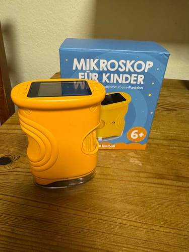 Rezensent zeigt das orangefarbene Abenteuer-Mikroskop auf einem Holztisch, mit der blauen Verpackung im Hintergrund.