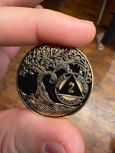 Customer holding the front of the black and gold 2 Year Sobriety Medallion, showing the Twisted Tree of Life design.