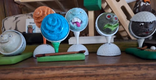 Reviewer's photo of several creative, hand-carved golf balls with funny faces sitting on tees on a shelf.
