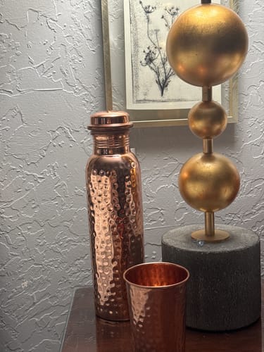 Reviewer's hammered Himalayan Ayurvedic Pure Copper Bottle and matching cup on a wooden surface.
