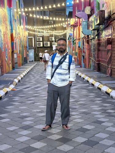 Reviewer standing in a colorful alley, wearing the grey 9 O'clock Pants with a casual shirt and sandals.