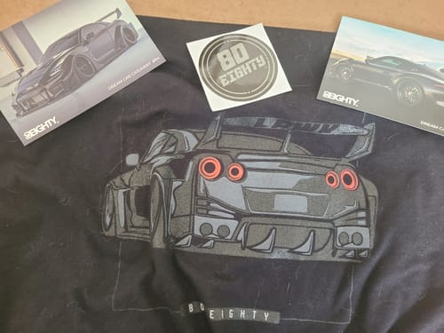 Customer's black graphic DCG84 Shirt showing the back of a car, laid flat with two cards and a sticker.