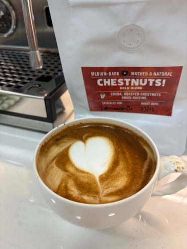 Customer's cup of coffee with heart-shaped latte art, with the ChestNuts! Bold Blend packaging in the background.