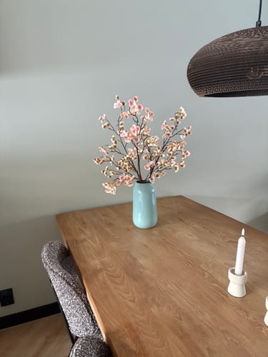 Klant toont de Oudroze Zijden Bloesems in een lichtblauwe vaas op een houten tafel.