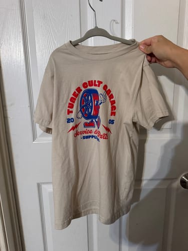 Reviewer holding the beige YOUTH WHEELIE DA WHEEL TEE on a hanger, showing the front graphic.