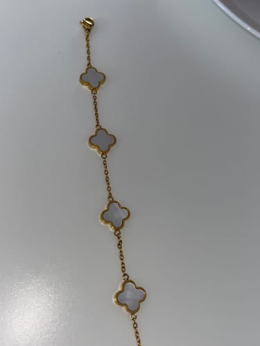 Rezensent zeigt das goldfarbene Kleeblatt-Armband mit vier weißen Anhängern, das auf einer weißen Oberfläche liegt.
