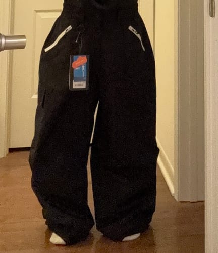 Reviewer standing indoors wearing the baggy black snow pants, with the product tag still attached.