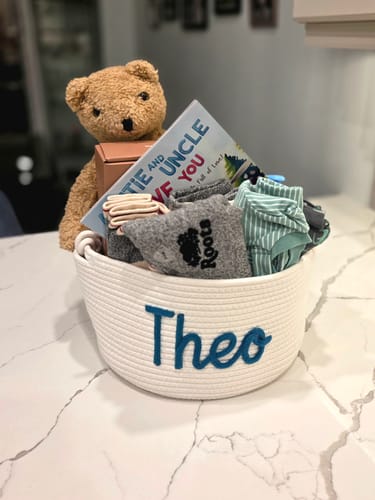 Customer's large white rope basket, personalized with 'Theo' in blue, holding a teddy bear, book, and clothing.