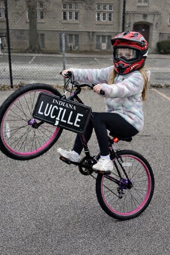 A person on a bicycle doing a wheelie with a personalized 'LUCILLE' Indiana Mini Blackout Metal License Plate on the front.