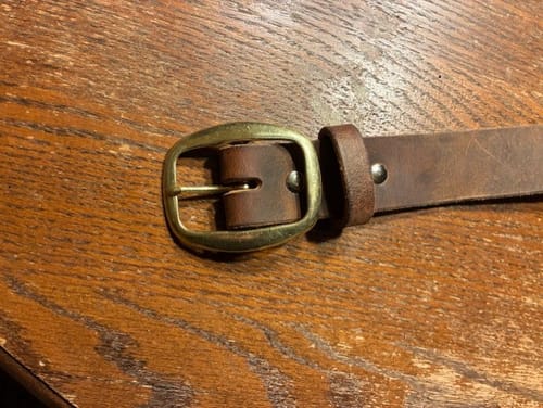 Reviewer's close-up of The Crazy Horse belt with a new solid brass buckle on a wooden surface.