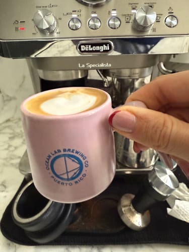 Customer's hand holding a small pink Ceramic Coffee Cup with frothed coffee in front of an espresso machine.