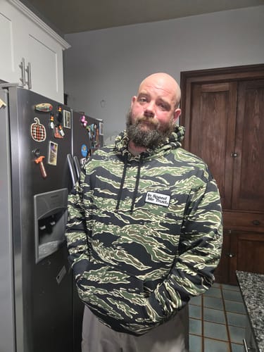 Reviewer standing in a kitchen wearing the TIGER CAMO Hoodie, showing the fit and camouflage pattern.