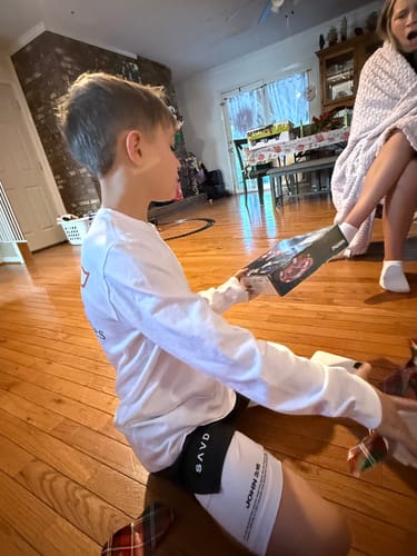 Customer sits on the floor wearing the white John 3:16 'The Cross' Youth Performance Shorts as an underlayer.