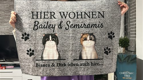 Ein Kunde hält die personalisierte graue 'Hier wohnt' Fußmatte mit zwei Katzenillustrationen und Namen hoch.