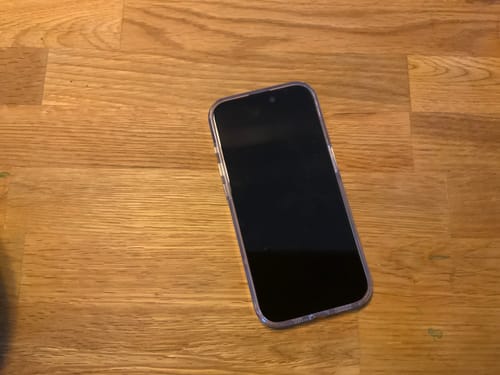 Ein Kunde zeigt sein iPhone mit der blasenfrei angebrachten 3-in-1-Schutzfolie, das auf einem Holztisch liegt.