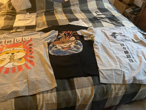 Reviewer's three graphic tees, including the Koi Harmony T-Shirt, laid out on a bed.