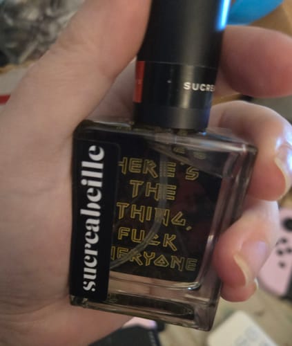 A reviewer's hand holds the square glass 'Here's the Thing, Fuck Everyone' perfume bottle.