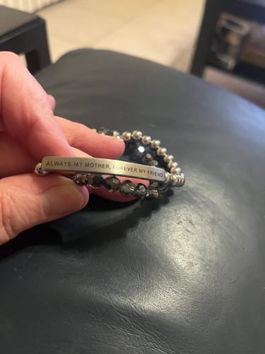 A customer holds the engraved 'Always My Mother, Forever My Friend' bracelet, shown stacked with a dark beaded bracelet.