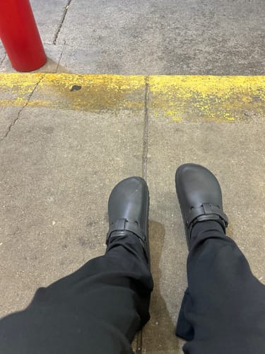 A first-person view of a reviewer wearing black The Cloudies Clogs while standing on a concrete sidewalk.