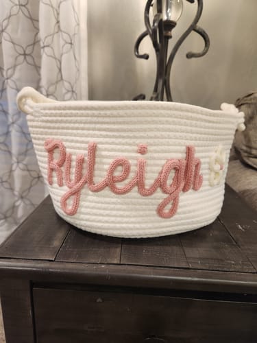 Customer's personalized white knitted basket with 'Ryleigh' in pink yarn and a flower icon, on a dark wooden table.