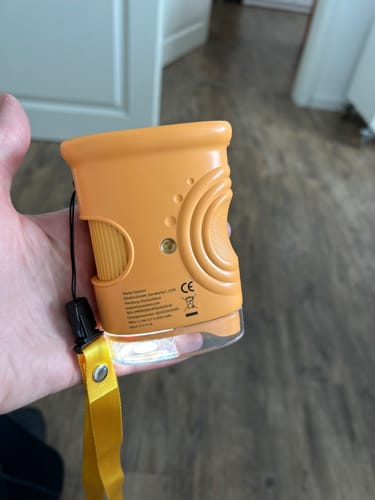 Ein Rezensent hält die orangefarbene Becherlupe des Edubini 'Gefunden!'-Spiels in der Hand, das Licht ist eingeschaltet.