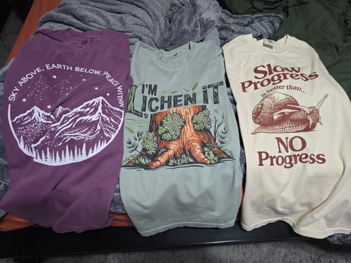 Customer's 'I'm Lichen It' T-shirt laid out on a bed between two other graphic tees.