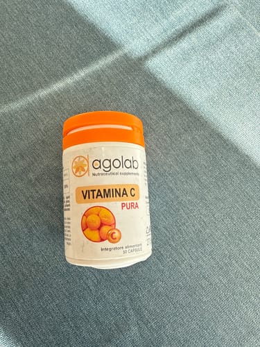 Recensore mostra il flacone bianco di Vitamina C Pura con tappo arancione su una superficie blu.