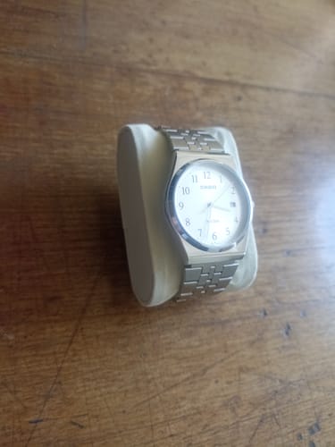 Cliente mostrando su Reloj Casio Análogo MTP-B145D-7BV plateado con esfera blanca, exhibido sobre una superficie de madera.