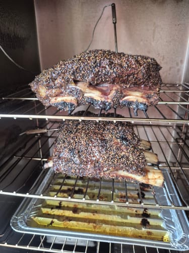 Customer's seasoned Bone-In Short Ribs cooking on two racks inside a smoker.