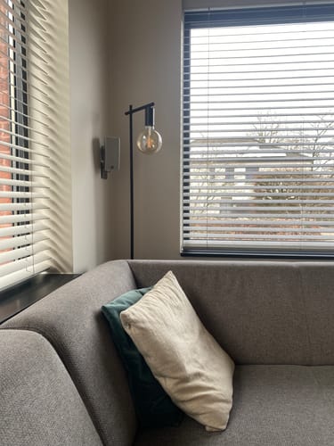 Recensent toont de zwarte Lucide LEANNE vloerlamp in een woonkamerhoek naast een grijze bank.