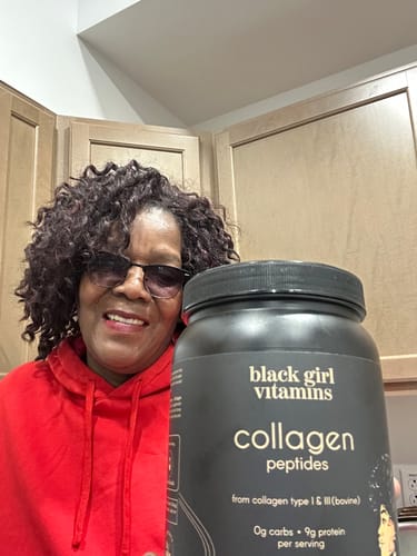 Reviewer smiling while holding a large black tub of BGV Collagen Powder in a kitchen.