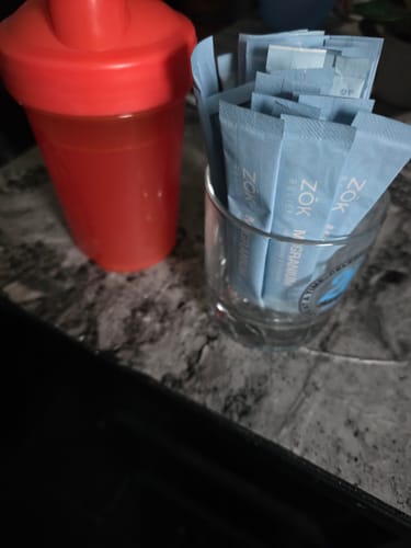 Customer's Migranium powder sachets in a glass next to a red shaker bottle on a kitchen counter.