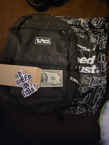 Customer's black "PATHWAY" BACKPACK laid out with a two-dollar bill, stickers, and branded bag it arrived with.
