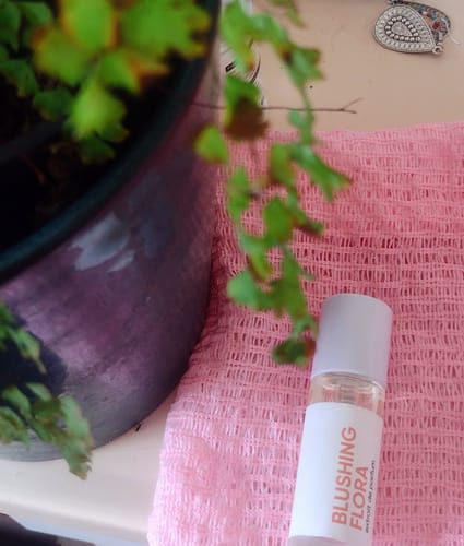 Customer's Blushing Flora perfume bottle resting on a textured pink cloth next to a plant.
