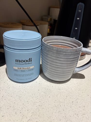 Customer's light blue tub of Moodi Mylk Chocolate blend next to a prepared drink in a grey, ribbed mug on a counter.