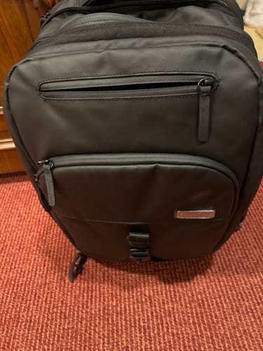 Customer's close-up of the black backpack, showing the front zippered compartments on a red carpet.