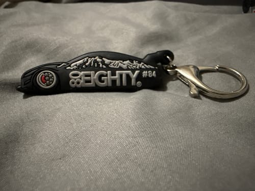 A customer's black 80Eighty DCG#84 keychain, shaped like a car, resting on a gray fabric surface.