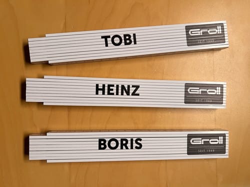 Ein Kunde zeigt drei personalisierte Zollstöcke mit den Namen Tobi, Heinz und Boris auf einer Holzoberfläche.
