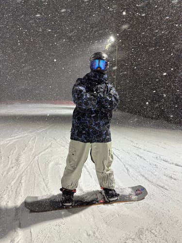 Reviewer wearing Men's Off White Snow Pants while standing on a snowboard on a snowy night.