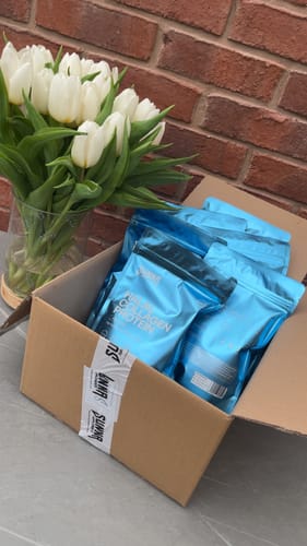 Customer's Black Friday Bundle of collagen protein pouches sitting in an open cardboard box next to a vase of white tulips.