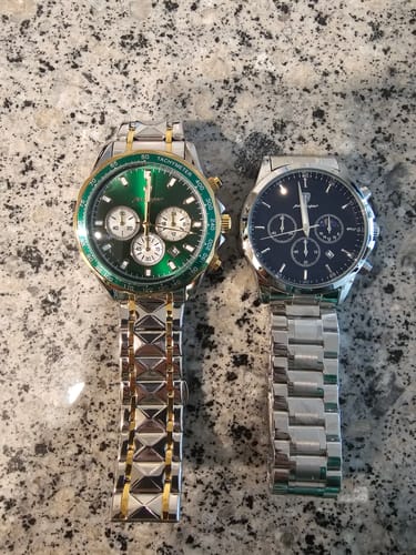 Reviewer's two watches on a stone countertop; one is two-tone with a green face, the other is silver-tone with a dark blue face.