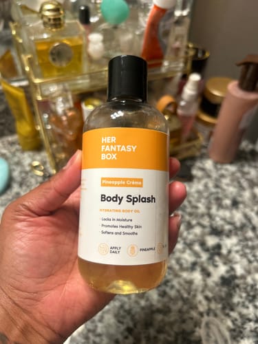 Reviewer holding a clear bottle of the Pineapple Crème Body Splash, showing its golden oil and orange label.
