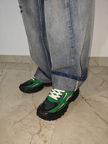 Close-up of a customer wearing the green and black COLOSSUS shoes with cuffed baggy jeans on a marble floor.