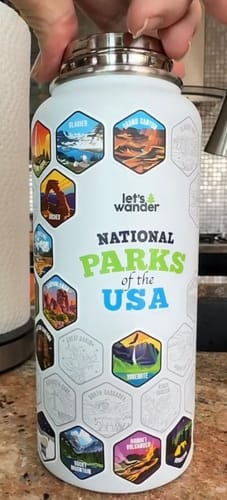 A reviewer's white National Parks Water Bottle, with colorful stickers applied over engraved outlines and a hand on the lid.