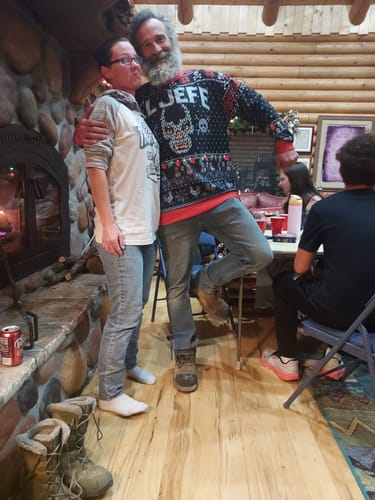 Customer posing in the black EL JEFE Navidad Sweatshirt with another person in a rustic-looking room.